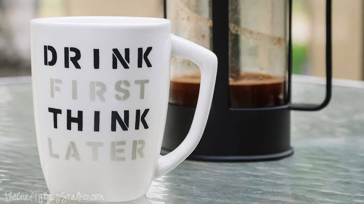 A white coffee cup with the words 'drink first, think later' inscribed in bold lettering, sitting on a glass surface beside a French press filled with coffee.