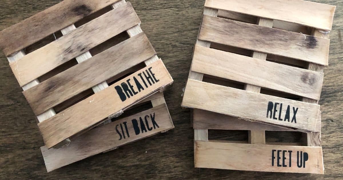 Two handcrafted mini pallet wood coasters featuring rustic slats, displaying stamped phrases like breathe, sit back, relax, and feet up on a wooden surface.