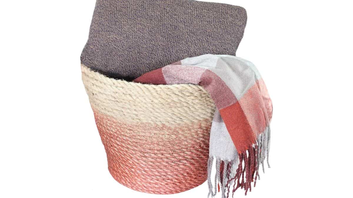 Woven twine basket housing a folded blanket within and a checked scarf cascading over the edge, styled as warm home decor.