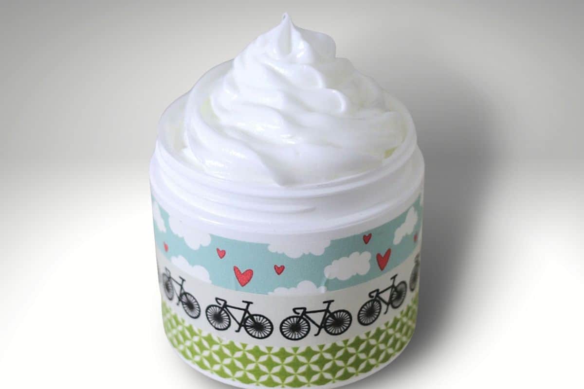 Open container of homemade whipped lotion featuring a silky, fluffy swirl atop and a decorative patterned label, captured against a light backdrop.