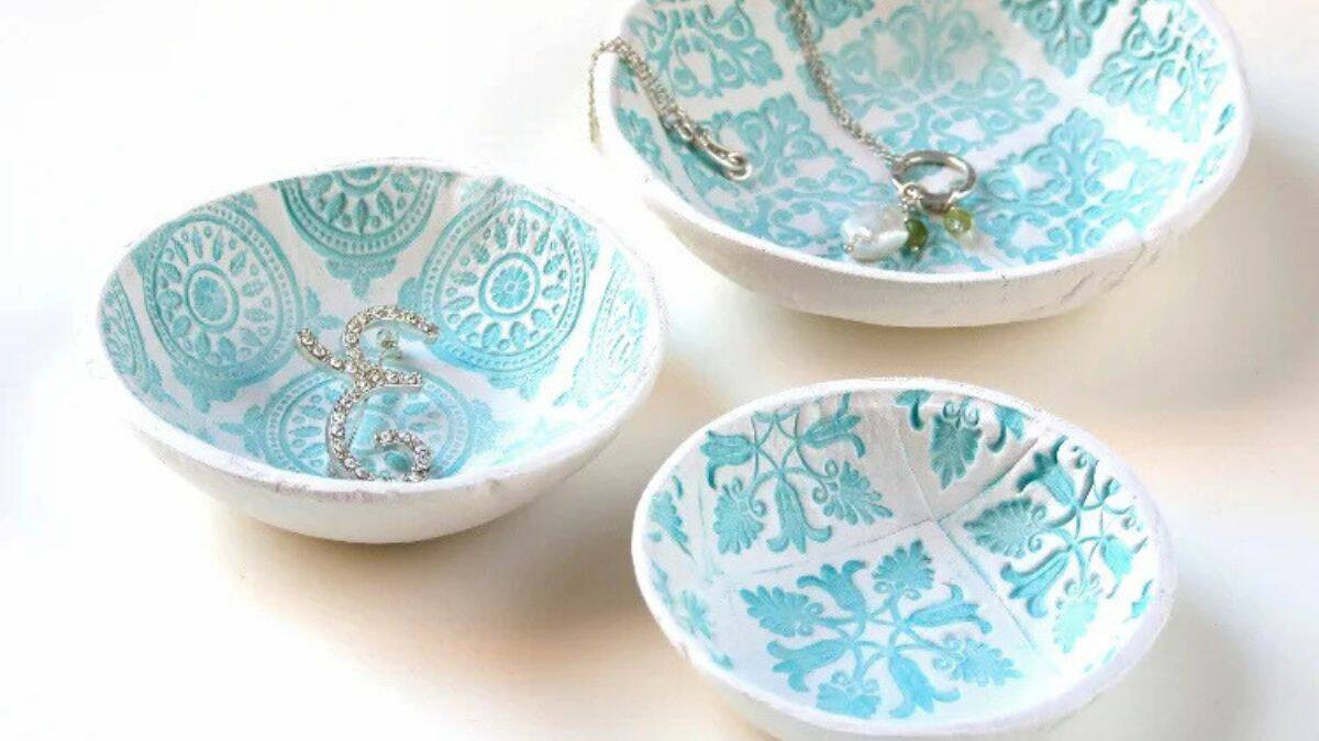 Three crafted stamped air-dry clay bowls featuring ornamental designs, arranged on a table holding small jewelry items such as rings and necklaces.