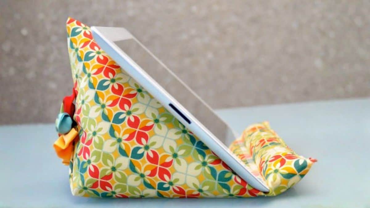 A crafted fabric iPad holder featuring a cushioned triangular design, supporting a tablet upright on a surface for hands-free viewing or reading.