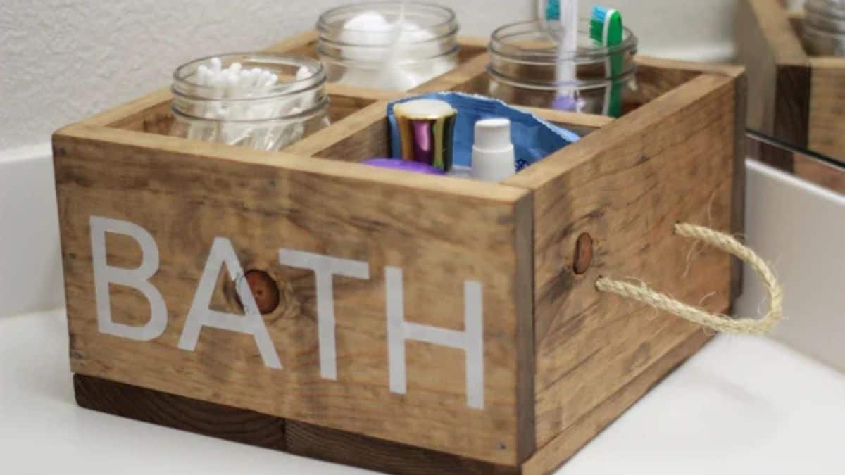 A wooden caddy for the bathroom with rope handles holding glass containers filled with cotton swabs, cotton balls, toothbrushes, and toiletries atop a bathroom counter.