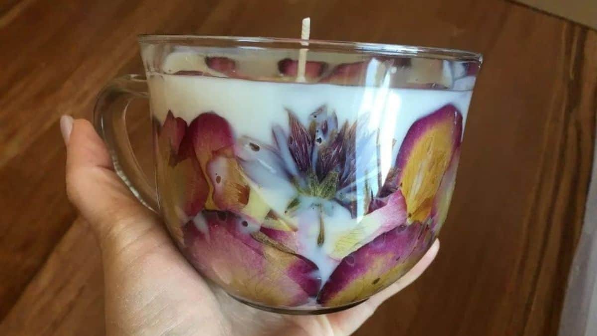 A hand grasping a transparent glass mug candle crafted with pressed flowers set in the wax and a central wick.