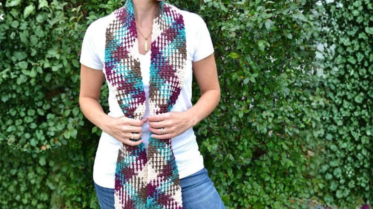 Individual sporting a handmade crochet scarf with color pooling, depicted draped around the neck and grasped at the ends while positioned outdoors in front of foliage.