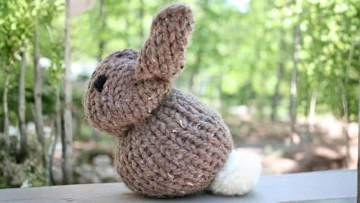 Knit stuffed bunny seen from the back, featuring floppy ears and a fluffy tail, resting on a wooden surface outdoors against a backdrop of trees.