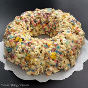 Ring-shaped popcorn cake composed of marshmallow-coated popcorn and candy sprinkles, shaped like a bundt cake and set on parchment paper.