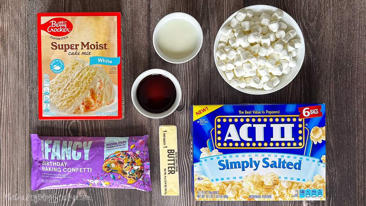 Ingredients for a popcorn cake displayed on a table, featuring cake mix, microwave popcorn, marshmallows, butter, vanilla, sweetened condensed milk, and confetti sprinkles.