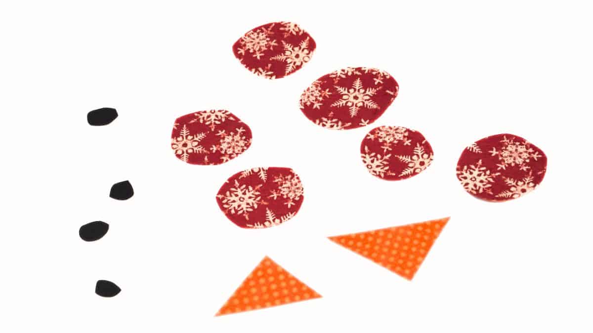 Paper shapes cut out for a snowman craft, including small black pieces for eyes and mouth, patterned circles for buttons, and triangular shapes for carrot noses.