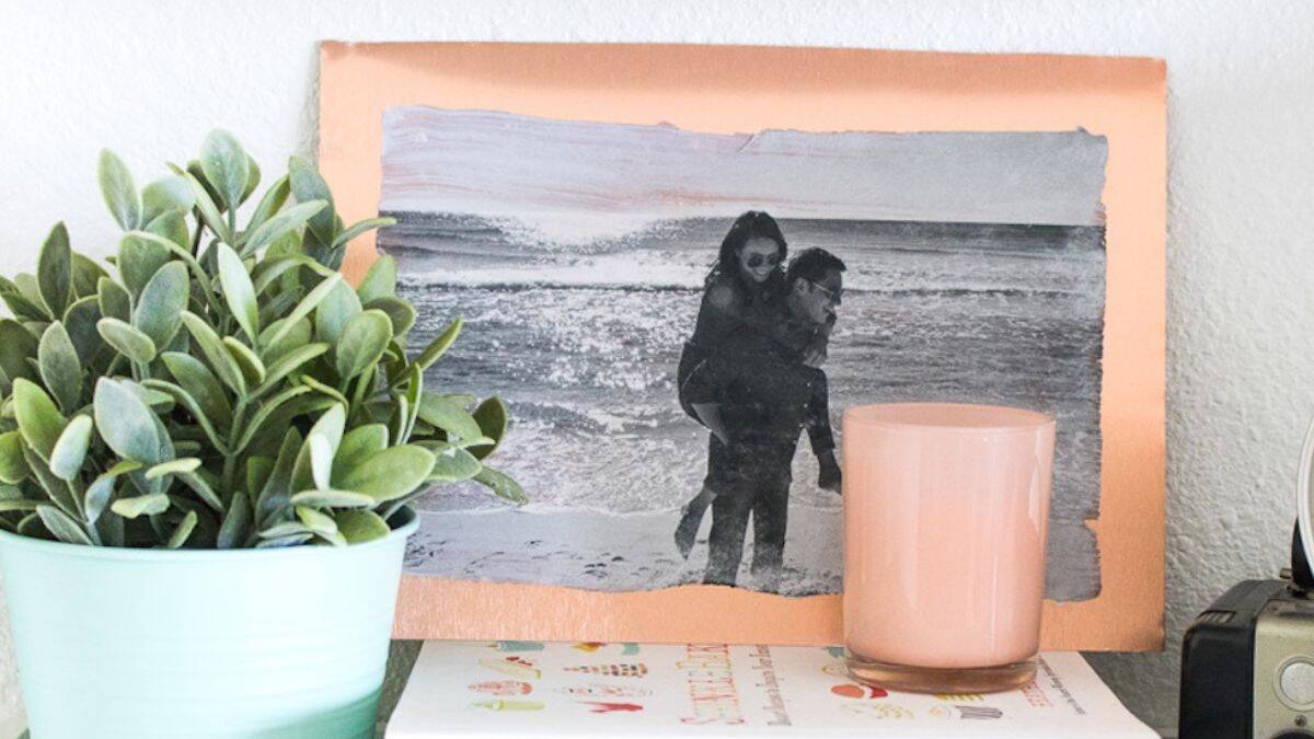 A metallic photo transfer of a duo at the shoreline exhibited on a shelf alongside a potted plant, candle, vintage camera, and stacked literature.