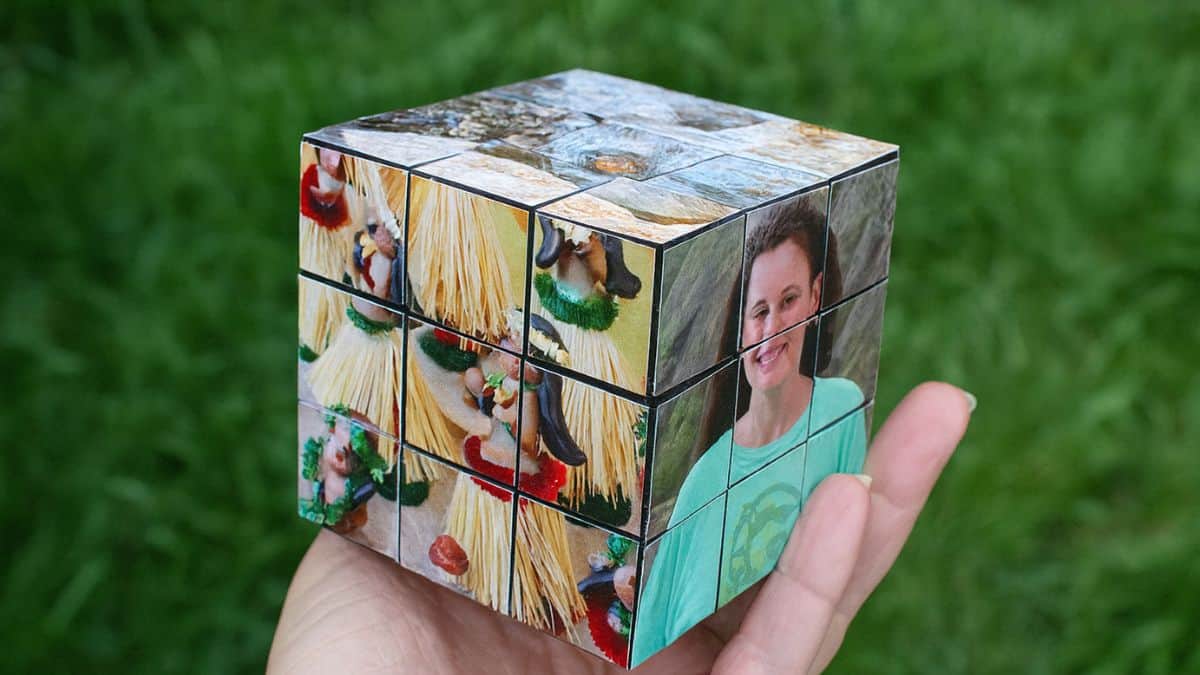 Hand grasping a customized photo cube formed from small square tiles, featuring family pictures arranged like a Rubik's cube set against a grassy backdrop.