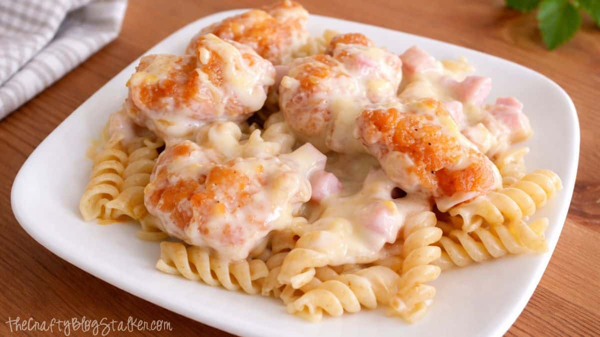 Cordon bleu casserole with chicken served over rotini pasta, garnished with crispy popcorn chicken, diced ham, and creamy melted cheese sauce on a plate.