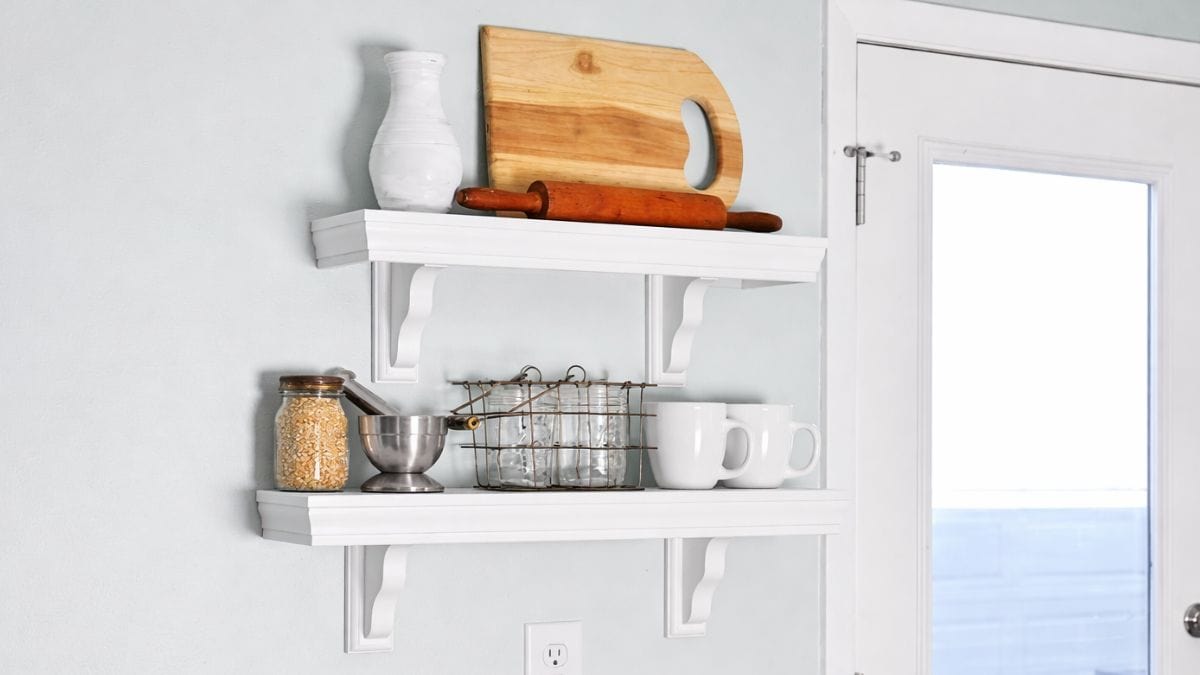White wall shelves adorned with a cutting board, rolling pin, mugs, jars, and kitchen paraphernalia in a farmhouse kitchen beside a glass door.