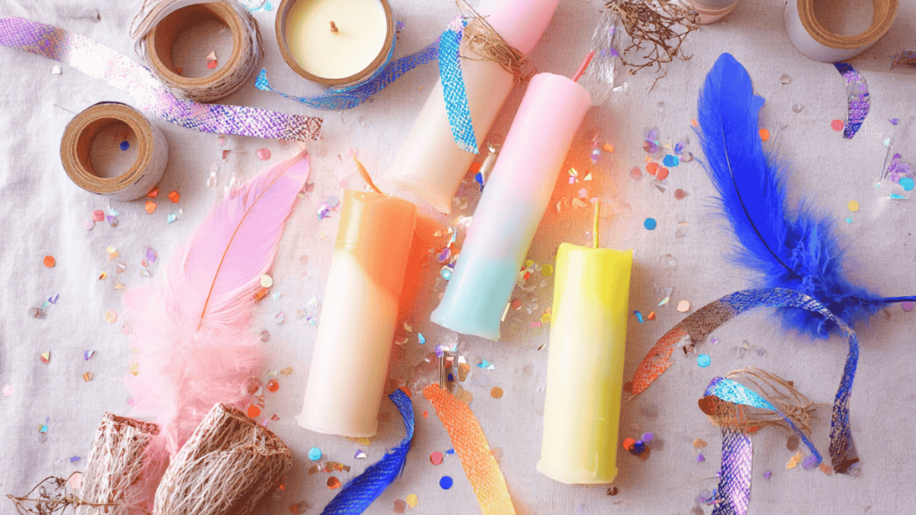 Candles using the dip-dye technique lie between feathers and confetti on a base made of beige fabric.