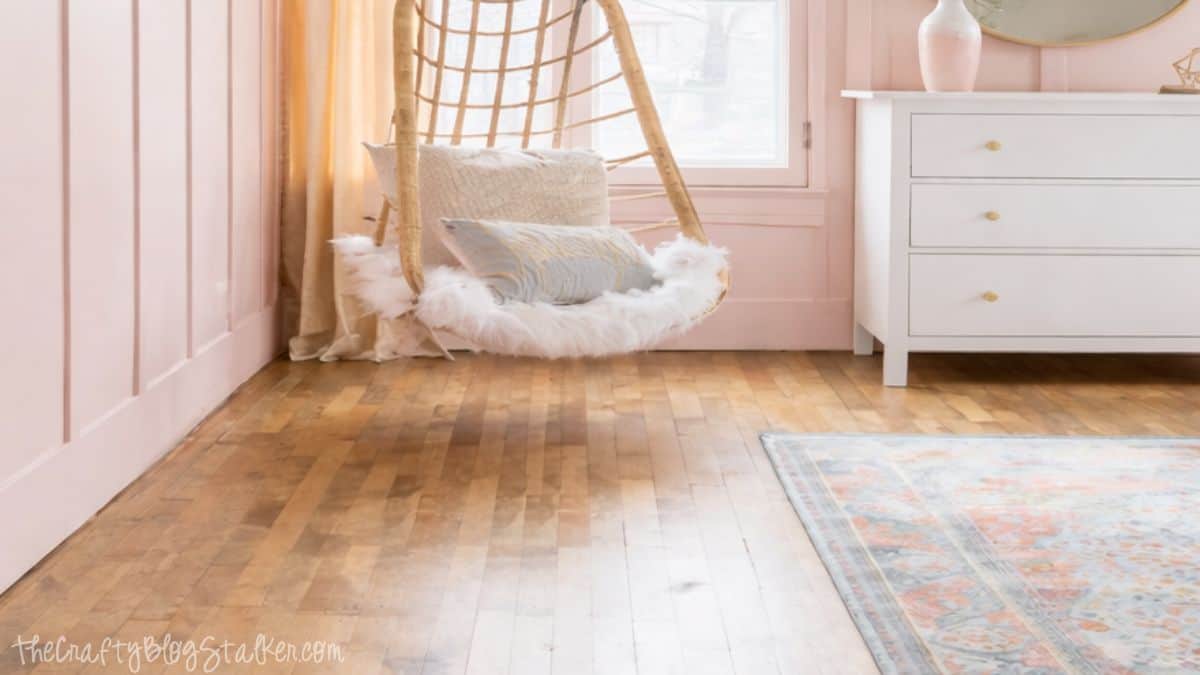 Hanging wicker chair with faux fur cushion and pillows near a window, adjacent to a white dresser on timber floors in a styled girl's bedroom.