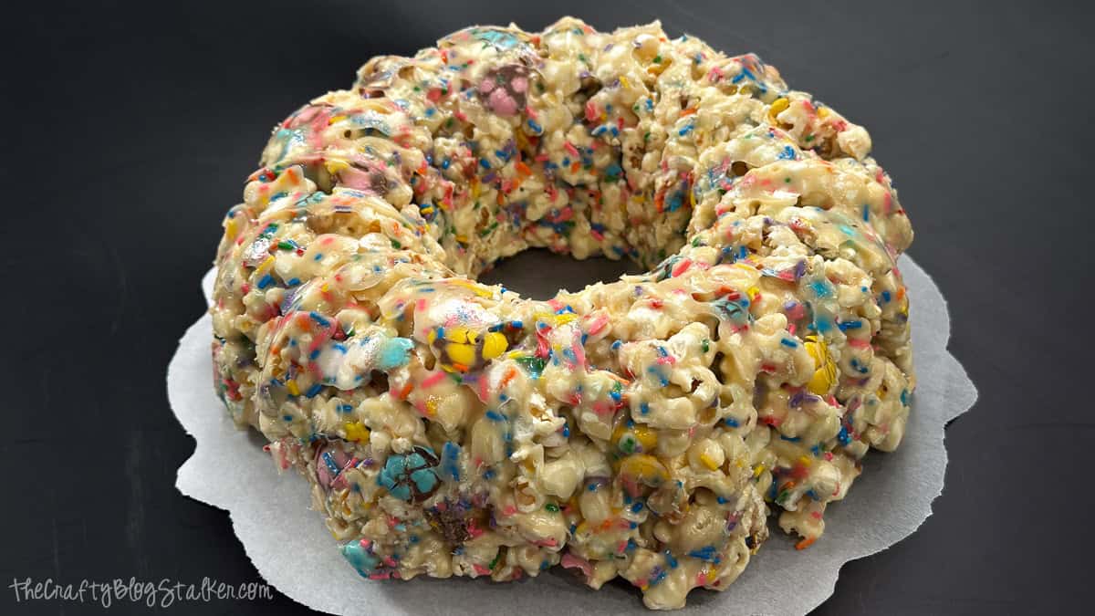 Ring-shaped popcorn cake crafted from marshmallow-coated popcorn with candy morsels and sprinkles, presented on parchment paper against a dark backdrop.