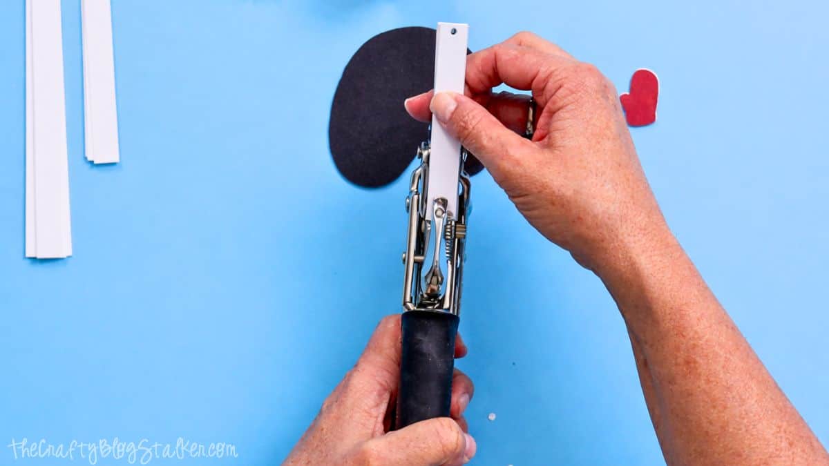 Hands utilizing a Crop-A-Dile tool to create a hole in a slim paper strip, with snowman craft components and paper strips on a crafting surface.