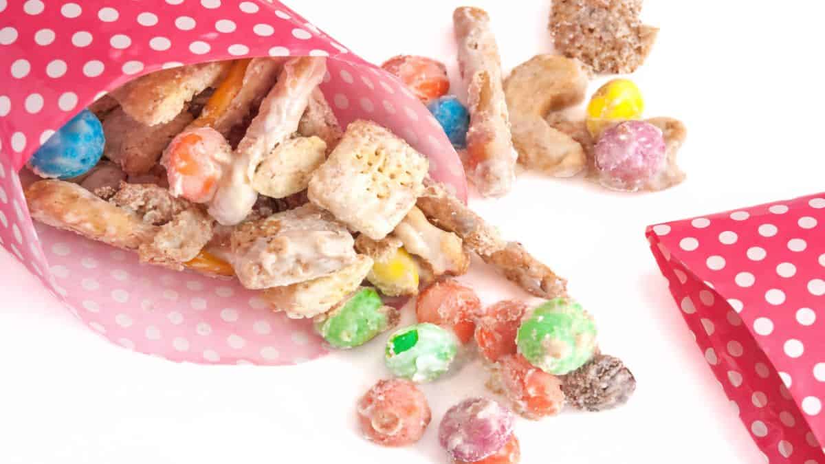 M&M White Chocolate Party Blend Recipe White chocolate party blend with cereal, pretzels, and sweets spilling from pink polka dot paper cones onto a white surface.